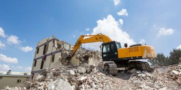 Yellow excavator demolishing a building under a bright blue sky.