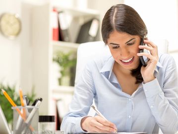A woman talking on phone while writing notes at her desk.