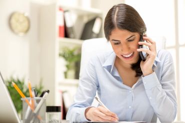 A woman talking on the phone while writing notes at her desk.