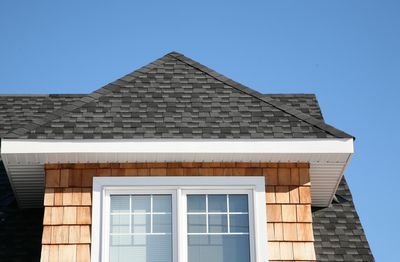 A rooftop with grey shingles and a window against a clear blue sky.
