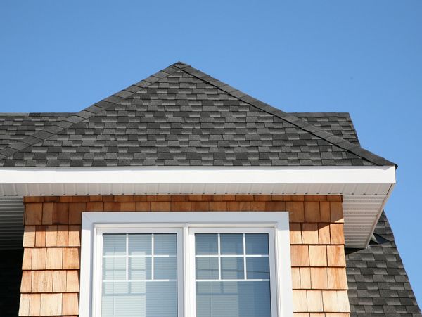House roof with grey shingles and wooden siding under a clear blue sky.