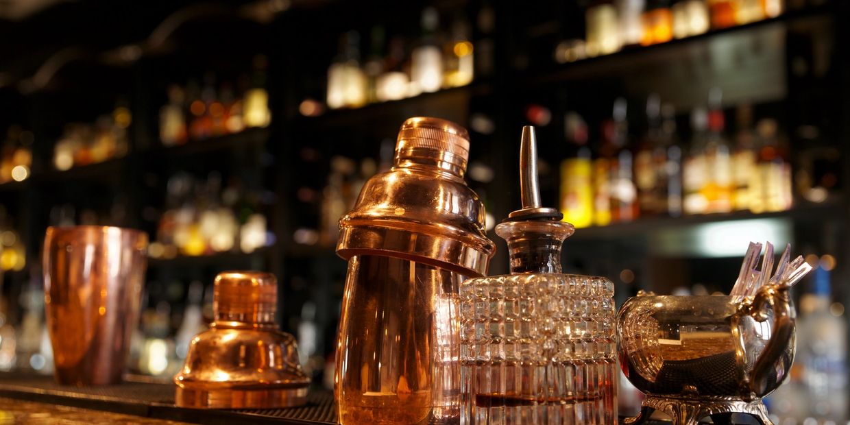 Copper cocktail shakers and glass decanter on a bar counter with blurred bottles in the background.