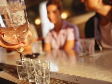 Bartender pouring shots into glasses at a bar with patrons in background.