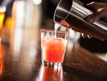 Bartender pouring a pink cocktail into a glass on a wooden bar.