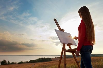 Artist painting on an easel with fields and lake in the background.