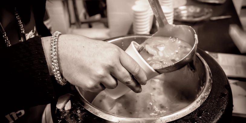 Hand ladling thick soup into a paper cup from a large pot.