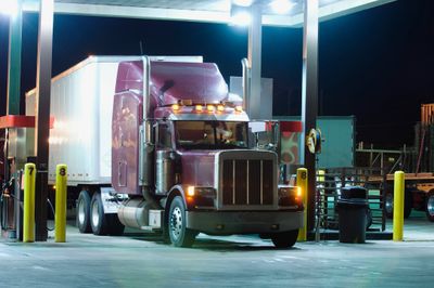 A maroon semi-truck refueling at a brightly lit gas station at night.
