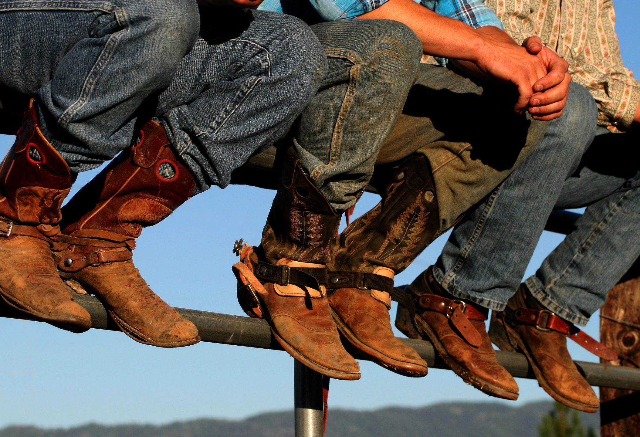 Cowboys sitting on a rail wearing worn boots and jeans under clear sky.