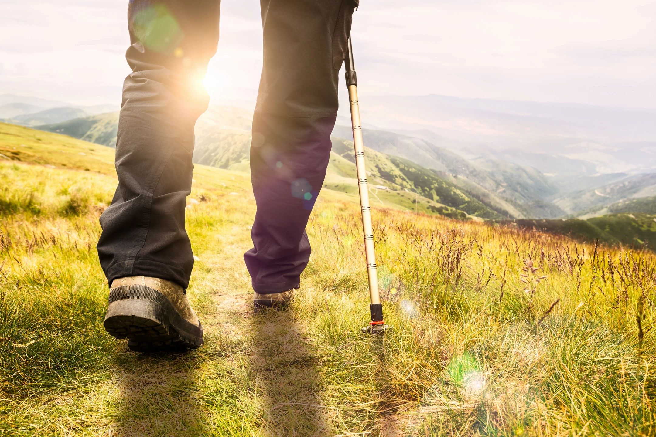 Legs walking toward a sunset over looking mountains while using a hiking pole.
