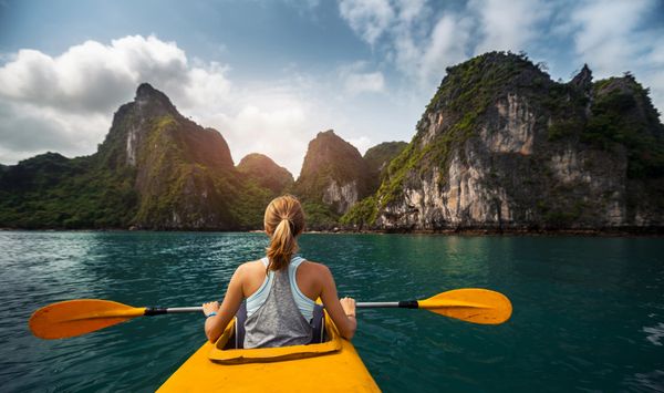 A woman kayaking towards lush, rocky islands on a calm sea under a partly cloudy sky.