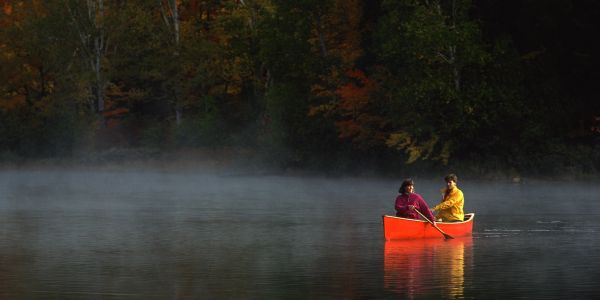 Two people in a bright orange canoe on a misty lake surrounded by autumn trees.
