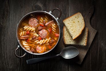 Hearty sausage and pasta stew served with slices of bread on a rustic wooden board.