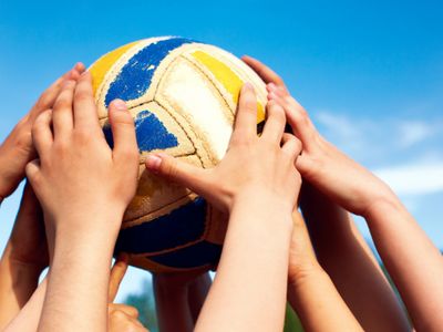 Multiple hands reaching for a volleyball against a clear blue sky.