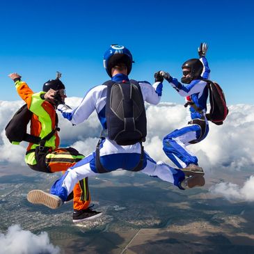 Three skydivers in colorful suits holding hands mid-air above clouds.