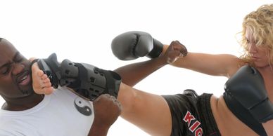 Woman in black gloves kicks man wearing a protective arm brace during martial arts training.