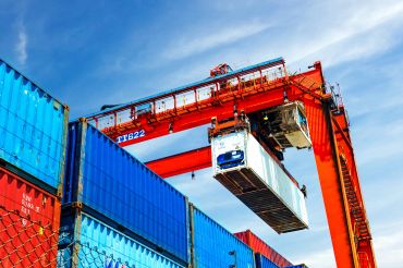 A large red crane lifting a shipping container at a busy port.
