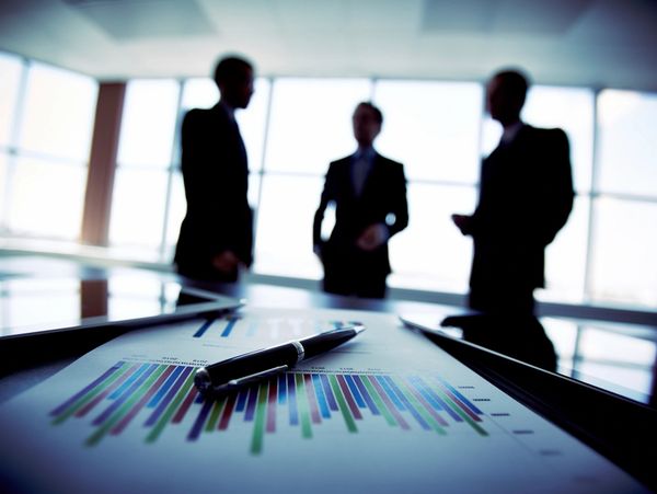 Business professionals discuss while charts and pen lie on table.