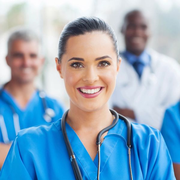 Smiling nurse in blue scrubs with stethoscope, colleagues in background.
