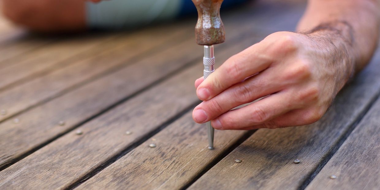 Hand using a hammer and punch tool on a wooden surface.