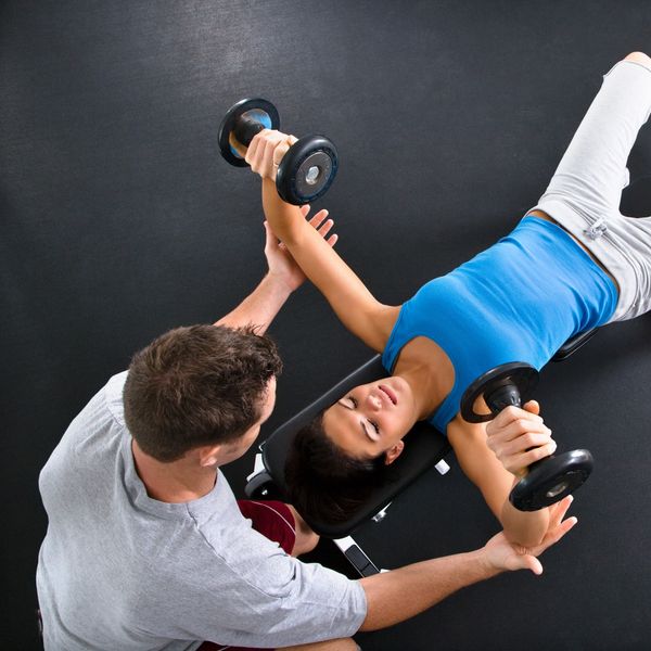 A woman lifts dumbbells with a trainer's assistance in a gym.