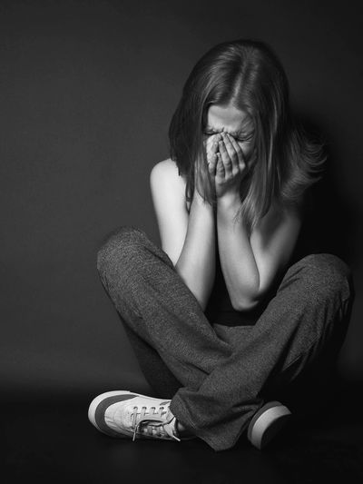 A woman sitting cross-legged, covering her face in distress against a dark background.