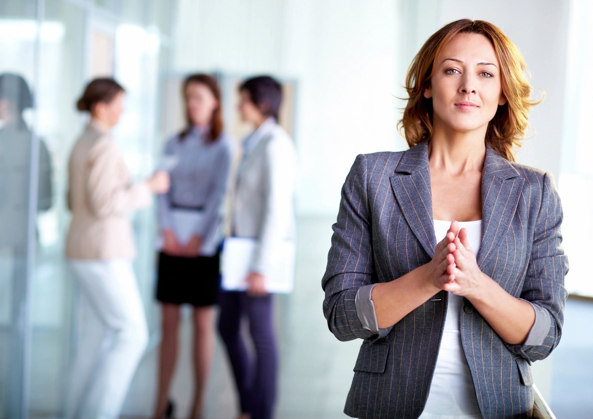 Confident businesswoman in a pinstripe blazer stands poised with colleagues blurred in the background.