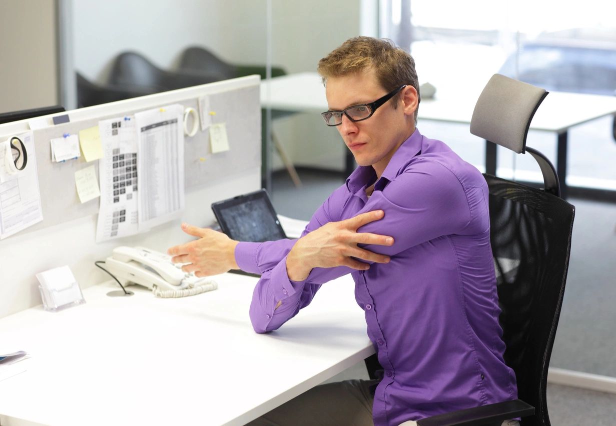 A worker stretches at his desk in his purple button up shirt.