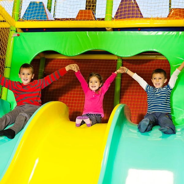 Three children joyfully slide down colorful slides holding hands indoors.