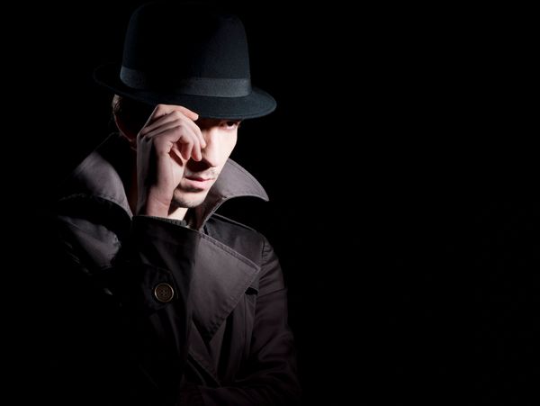 Man in trench coat and fedora hat in dramatic low light.