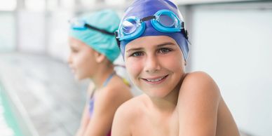 Two kids sitting at the side of a swimming pool, one child smiling.