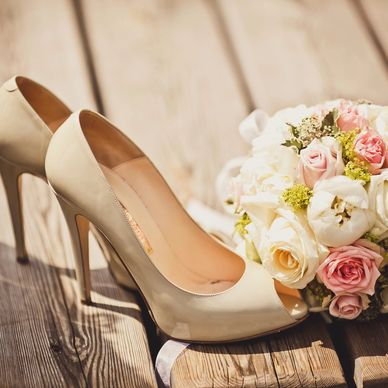 Elegant beige high heels next to a bouquet of pink and white roses on wooden floor.
