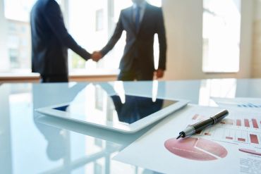 Two businessmen shaking hands behind a tablet and documents.