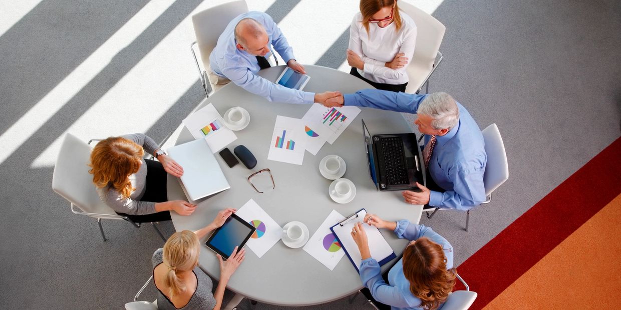 Top-down view of a table with a group of people working, two people reach across to shake hands