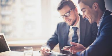 A consultant and client in suits conversing whilst sat at a table with laptop and paperwork.