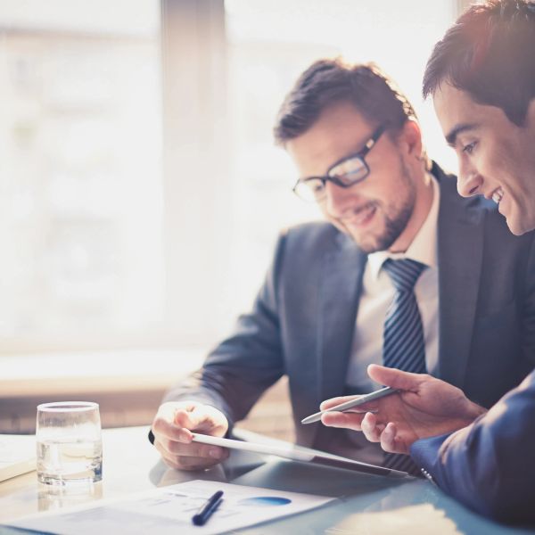 Two businessmen in suits discussing work over a tablet in a bright office.