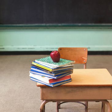 A stack of books with a red apple on top on a wooden school desk.