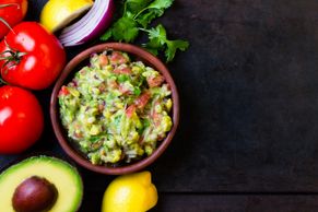 Fresh guacamole in a bowl surrounded by tomatoes, avocado, lemon, onion, and cilantro.