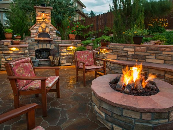 Cozy outdoor patio with stone fireplace and fire pit surrounded by cushioned chairs.