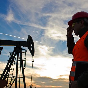 Worker in safety gear observes oil pumpjack against sunset sky.
