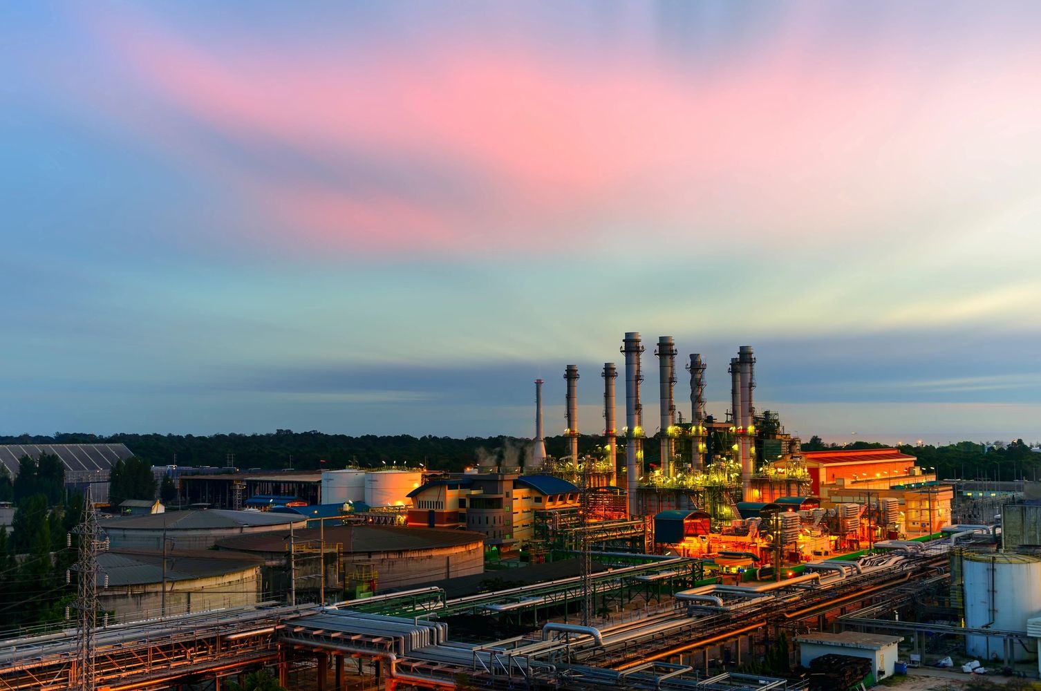 Image of a oil refinery in the early morning, with golden lights and a blue-pink clouded sky.