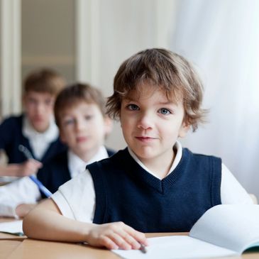Young boy in school uniform sitting at desk with open book, classmates blurred behind.