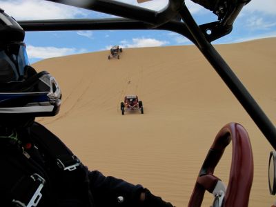 A person driving an off-road vehicle in the desert with others climbing a sand dune.