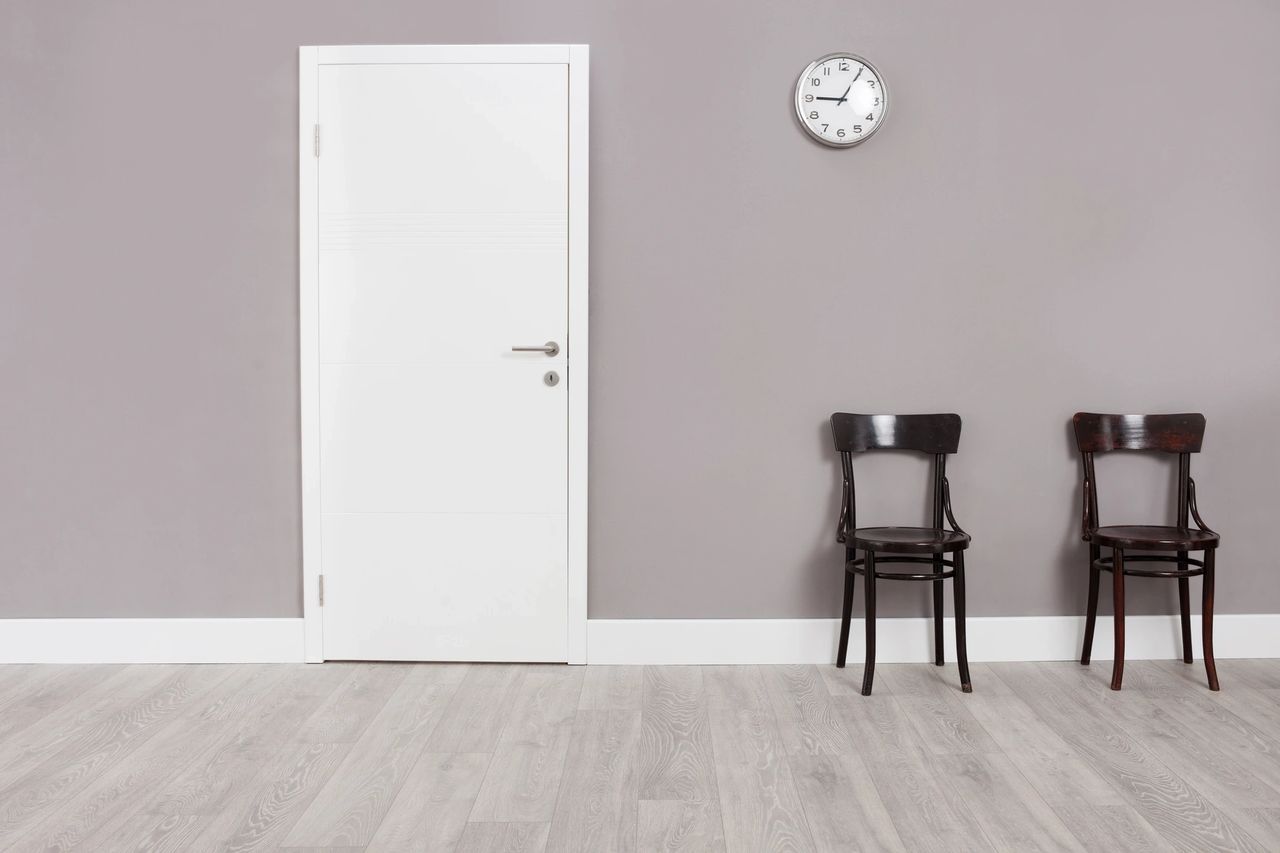 An empty room has two chairs siting against a wall, next to a door and a clock on the wall above them.