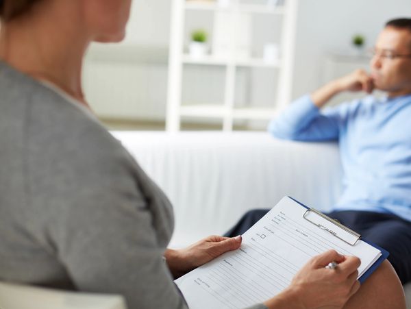 Therapist holding a clipboard listens to a man on a couch during a session.
