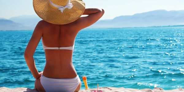 Woman in a white bikini and sunhat sitting by the sea, enjoying the sunny day.