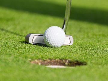 Close-up of a golf ball and putter near the hole on green grass.