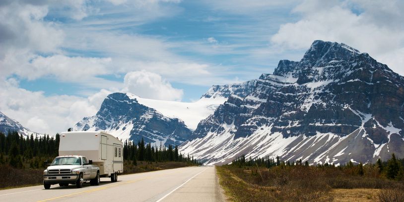 A truck towing a camper on a highway with snowy mountains and blue sky in the background.