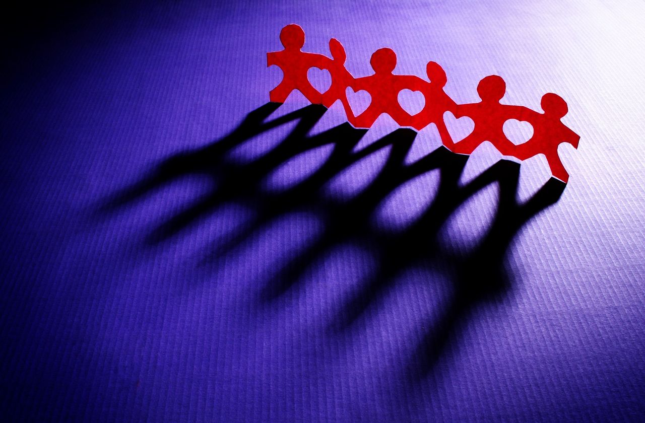 Origami of six red figurines of people holding hands stand on top of a purple backdrop.