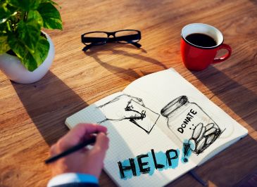 A person sketches a donation jar and the word HELP on a notebook with coffee and glasses nearby.