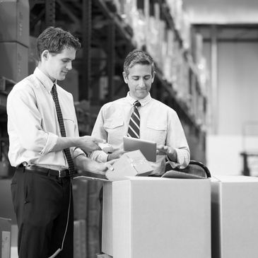 Two men in ties scanning packages in a warehouse with shelves of boxes.